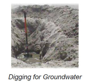 The image shows a deep, narrow pit dug into sandy soil, with sand piled up around its edges. The hole appears to have small amounts of water at the bottom, indicating moist or wet conditions. A slender, vertical tool or implement with a black and red handle is positioned within the pit, suggesting an ongoing process of digging or probing for water. The surrounding area is barren without visible vegetation or other features.