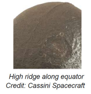 The image depicts a portion of Iapetus, one of Saturn's moons, showcasing its contrasting surface. The surface appears to have a dual-tone effect with a dark chocolate-brown area dominating the left side, gradually transitioning to a lighter, grayish region on the right. The dark part is marked by subtle, linear ridges and patterns that crisscross the surface. There are numerous craters visible, especially concentrated towards the center and the lighter region, indicating impact sites. The craters vary in size and are ring-like in appearance. The overall texture of the moon's surface appears rough and rugged.