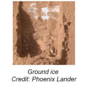 The image shows a rectangular trench on the surface of Mars, created by the Phoenix Mars Lander. The trench reveals a distinct layer of light-colored ground ice beneath the reddish, dusty Martian soil. The soil appears to have a coarse texture with small clumps and scattered rocks. The edges of the trench are uneven, with visible shadows highlighting the depth of the excavation. The composition focuses on the contrast between the reddish soil and the white ice layer, emphasizing the discovery made by the lander.
