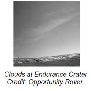 The image shows a grayscale landscape on Mars. The scene captures a wide expanse of sky and a rocky terrain in the foreground. The sky is mostly blank with a few thin, wispy clouds stretching diagonally across the image from the left towards the upper right. The terrain appears rugged and uneven, with a scattering of rocks and small peaks spread throughout the surface, giving it a textured appearance.